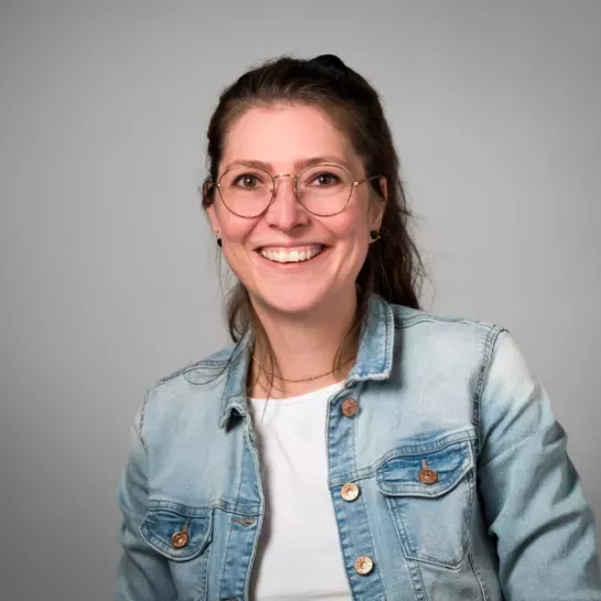 Eine Frau mit braunen Haaren und Brille lacht in die Kamera. Sie trägt eine Jeansjacke.