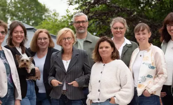 Vor dem Gabäude der Schulassistenz steht das Team, welches aus Männern und Frauen betseht. SIe sind alle schick gekleidet und richten ihren Blick in die Kamera.
