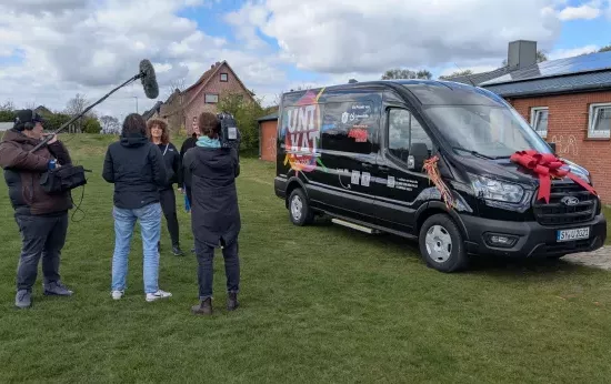 Ein Fernsehteam filmt die beiden Trainerinnen der Trommelgruppe. Daneben steht der neue Bus.
