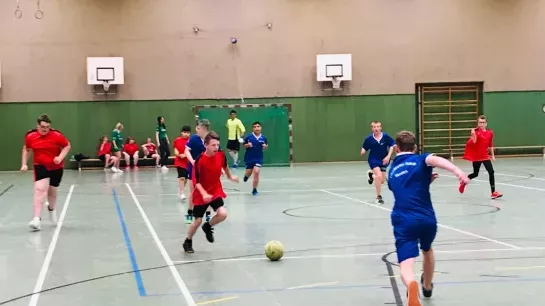 In einer Turnhalle spielen zwei Teams Fußball. Die eine Mannschaft trägt blaue und eine rote Trikots. Alle sind in Bewegung.