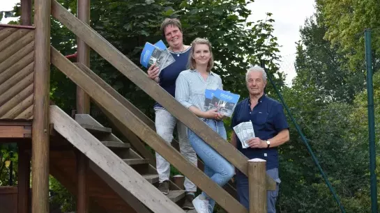 Zwei Frauen und ein Mann stehen auf einer Treppe eines Klettergerüstes und halten Bücher in ihren Händen. Sie stehen leicht schräg und richten ihren Blick lächelnd zur Kamera.