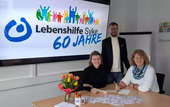 Zwei Frauen sitzen an einem Tisch und lachen in die Kamera. Hinter ihnen steht ein Mann. Im Hintergrund sieht man das Logo zu 60 Jahren Lebenshilfe Syke.