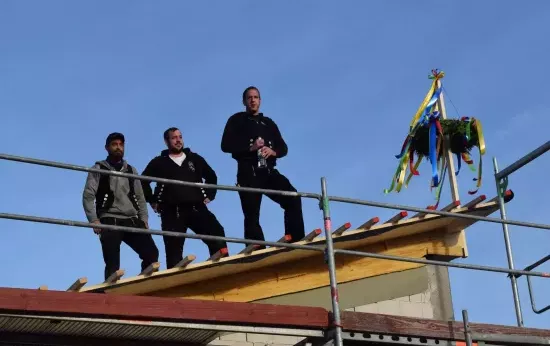 Drei Dachdecker stehen auf einem Dach. Einer hält eine Flasche in der Hand. Daneben ist ein Richtkranz am Dach festgemacht.