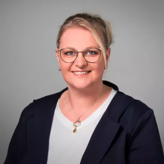 Eine blonde Frau mit Brille lächelt in die Kamera. Sie trägt einen blauen Blazer.