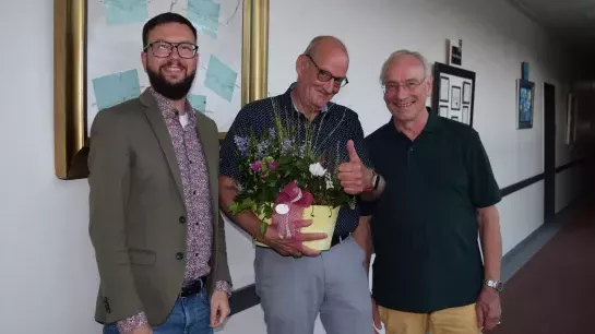 Geschenkübergabe. Drei Männer stehen in einem langen Flur, mit Bildern an den Wänden. Alle richten einen freundlichen Blick in die Kamera. Der Mann in der Mitte hält eine Schale mit Blumen in seiner Hand.