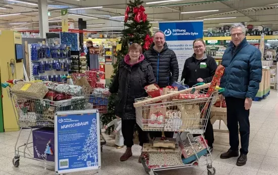 Zwei Frauen und zwei Männer stehen vor einem Weihnachtsbaum. Daneben stehen drei Einkaufswagen voll mit Geschenken.