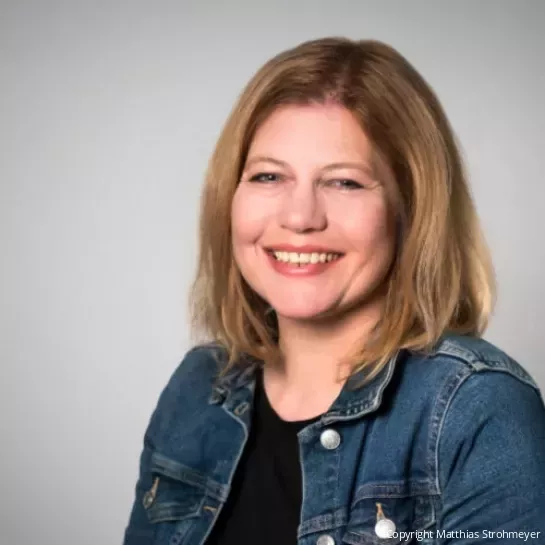 Yvonne Stiens, bei der Lebenshilfe Syke zuständig für Öffentlichkeitsarbeit uns Fundraising, sitzt vor einem grauen Hintergrund. Sie trägt eine Jeansjacke.