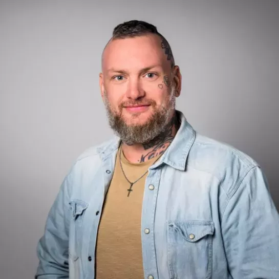 Raffael Saegler, Einrichtungsleitung Wohnheim Bassu, sitzt vor einem grauen Hintergrund. Er trägt ein Jeanshemd, hat einen Bart und einige Tattoos am Hals sowie links am Kopf.