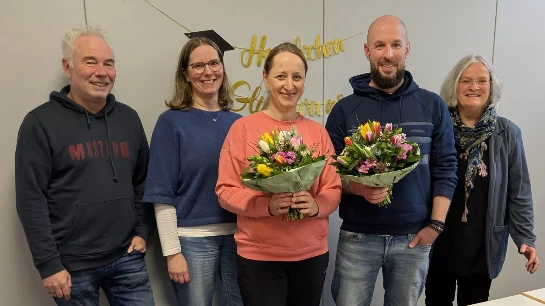 Abschluss Studium In der Mitte stehen ein Mann und eine Frau mit einem Blumenstrauß. Rechts und links davon stehen noch zwei Frauen und zwei Männer. Alle lächeln.