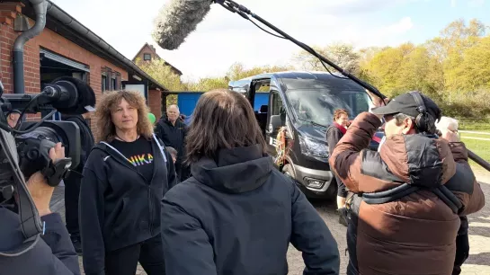 Eine Frau wird von einem Kamerateam interviewt. Im Hintergrund sieht man Menschen und einen Bus.