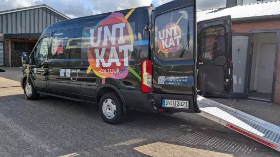 Ein schwarzer Bus mit bunten Logos hat die Türen hinten geöffnet. Eine Rollstuhlrampe ist ausgeklappt.