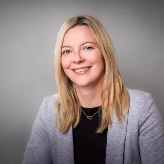 Eine blonde Frau mit langen Haaren lächelt in die Kamera. SIe tägt einen hellgrauen Blazer.